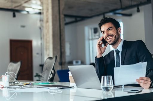 . A man in a suit sitting at a desk with a laptop and phone.and looking happy to talk with customer