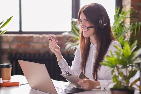 A woman with a headset and clipboard sits at a desk, working on a laptop.