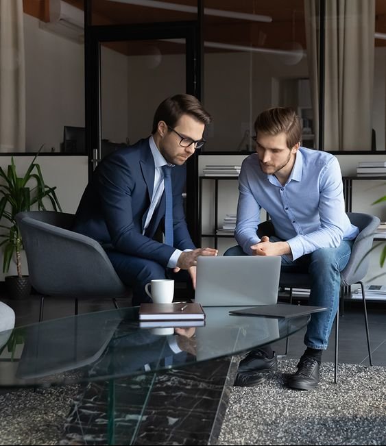 Two businessmen in suits sitting on a couch, focused on a laptop screen.